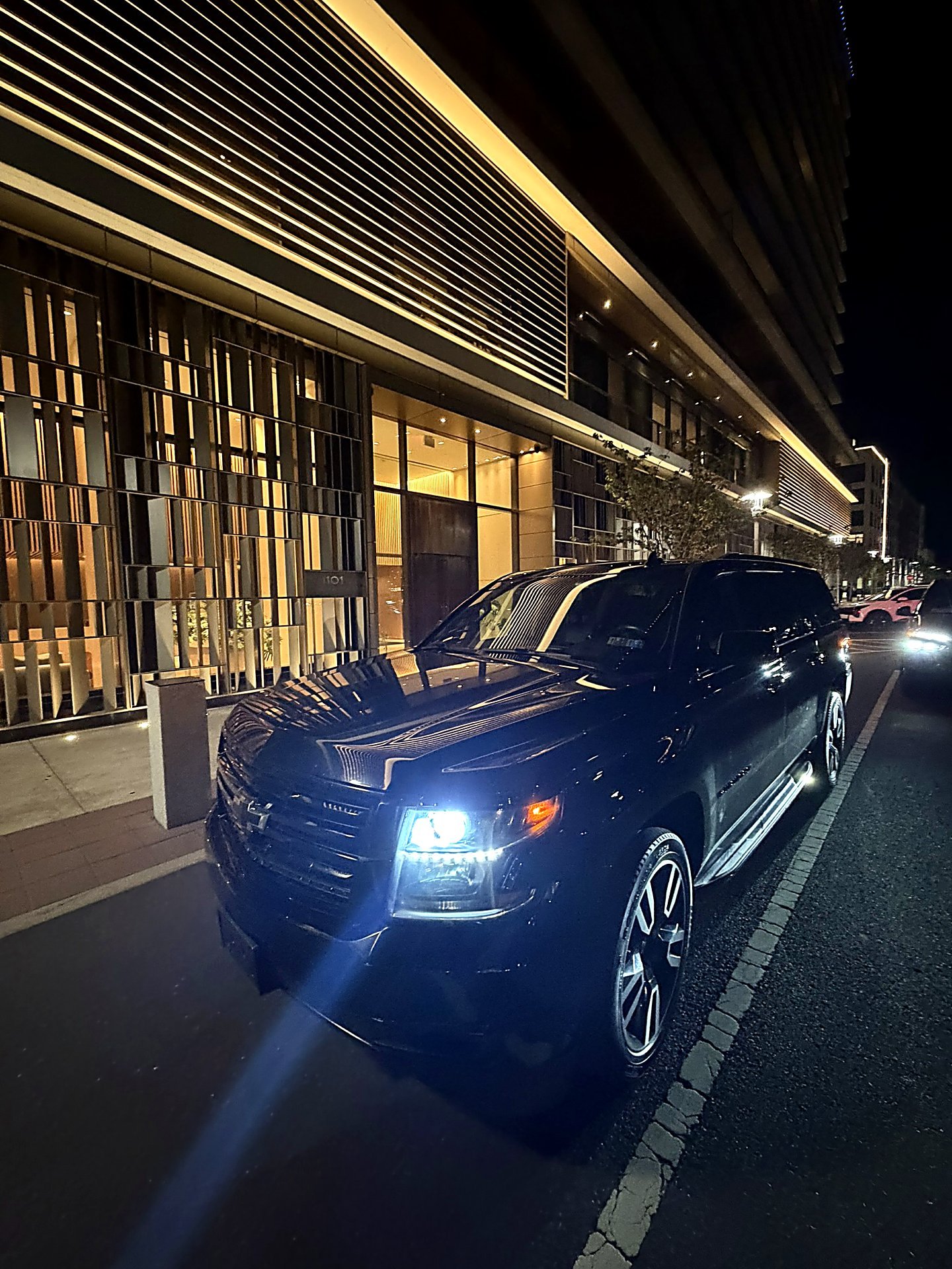 Black SUV with headlights on outside a modern building at night